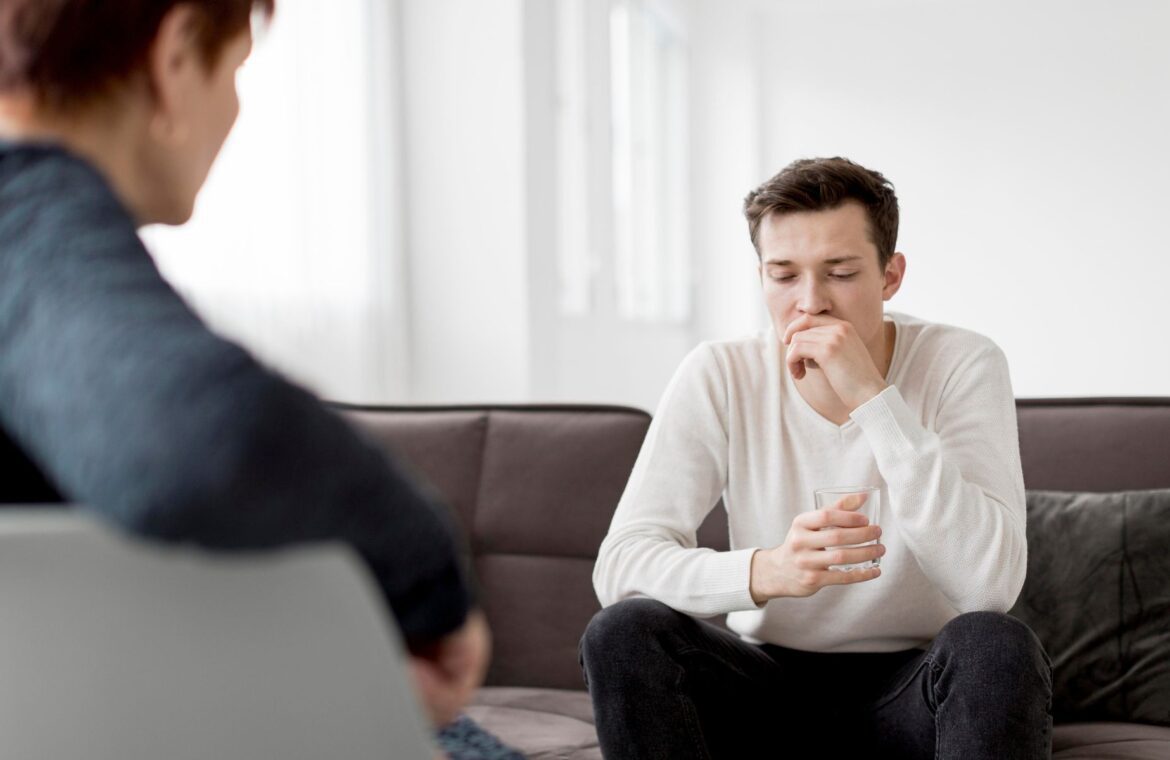 Psihoterapija – mlad moški sedi v naslanjaču in se pogovarja s psihoterapevtko v mirnem terapevtskem okolju.